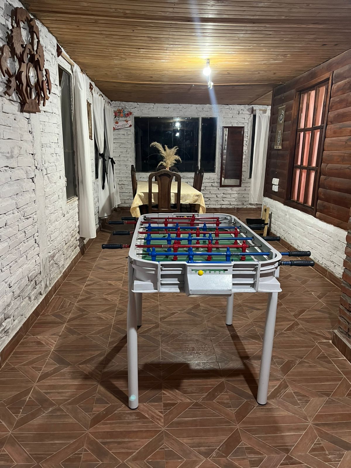A foosball table is positioned centrally in a well-lit room with wooden ceiling and tiled flooring. The walls are decorated with a mix of rustic wood and white-painted bricks. A dining table with chairs is visible in the background, along with window frames allowing natural light to enter.