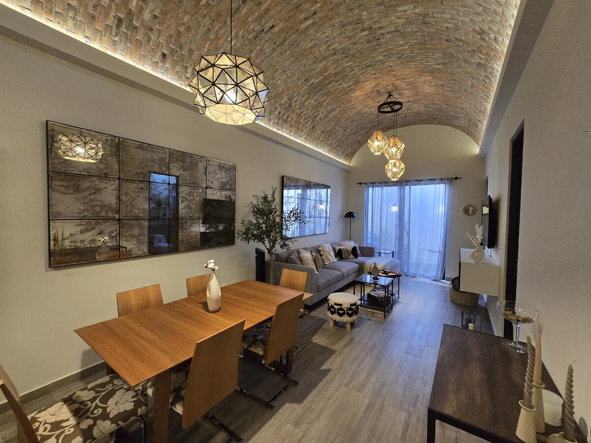 A spacious living area features a high, vaulted brick ceiling, illuminated by modern pendant lights. A wooden dining table sits beside a comfortable sofa, with soft lighting enhancing the cozy ambiance. Large windows provide natural light, leading to an outdoor space.