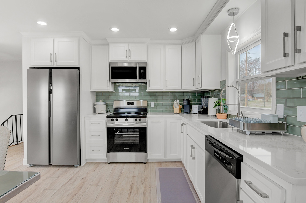 A modern kitchen features stainless steel appliances, including a refrigerator and oven. White cabinets complement the light-colored countertops, and a backsplash of green tiles adds a subtle splash of color. Natural light is invited through a window above the sink, with a dishwasher integrated into the cabinetry.