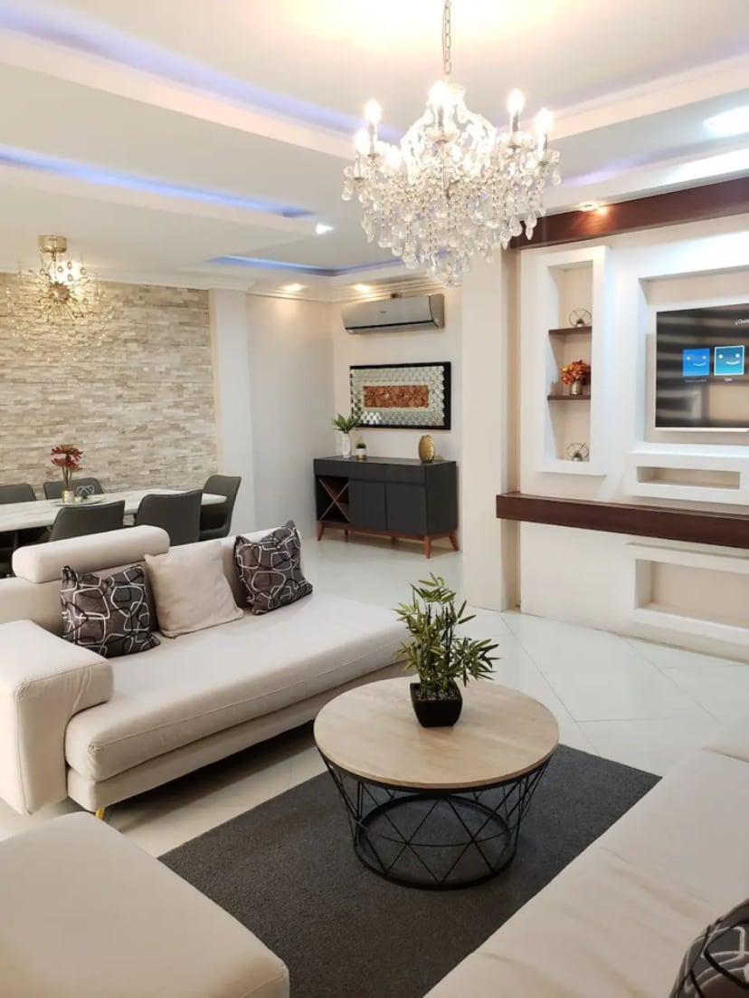 A modern living area is featured, showcasing soft beige sofas paired with patterned cushions. A round coffee table sits in the center, accompanied by a small potted plant. Elegant lighting is provided by a chandelier, and a decorative wall with shelving adds visual interest.