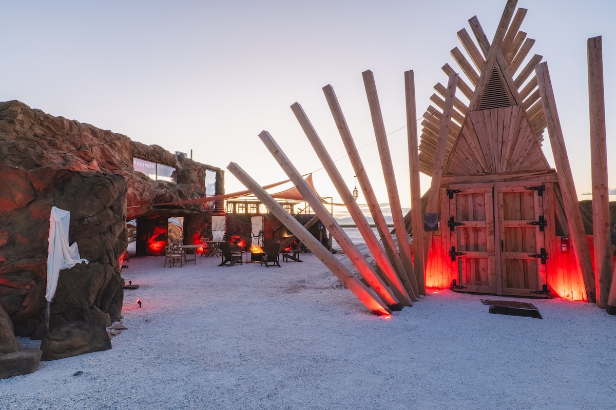 A uniquely designed structure stands against a twilight sky, featuring prominent wooden beams arranged in a striking pattern. The entrance showcases a large wooden door, while soft red lighting highlights the area. Surrounding elements include rugged stone textures and an open space with seating arrangements.