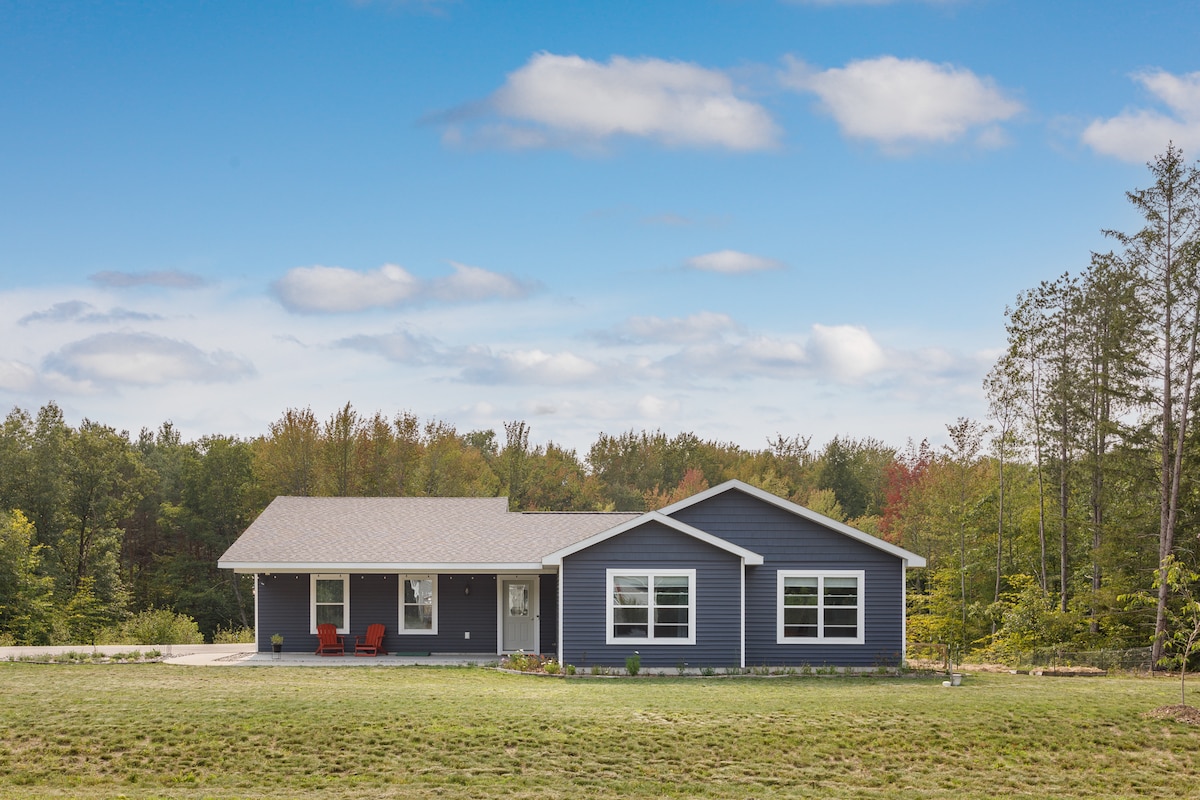 A modern home is showcased, with a dark blue exterior and multiple large windows. Red accent chairs are positioned on the porch, surrounded by a spacious green yard. A backdrop of trees and a clear blue sky enhances the peaceful setting.