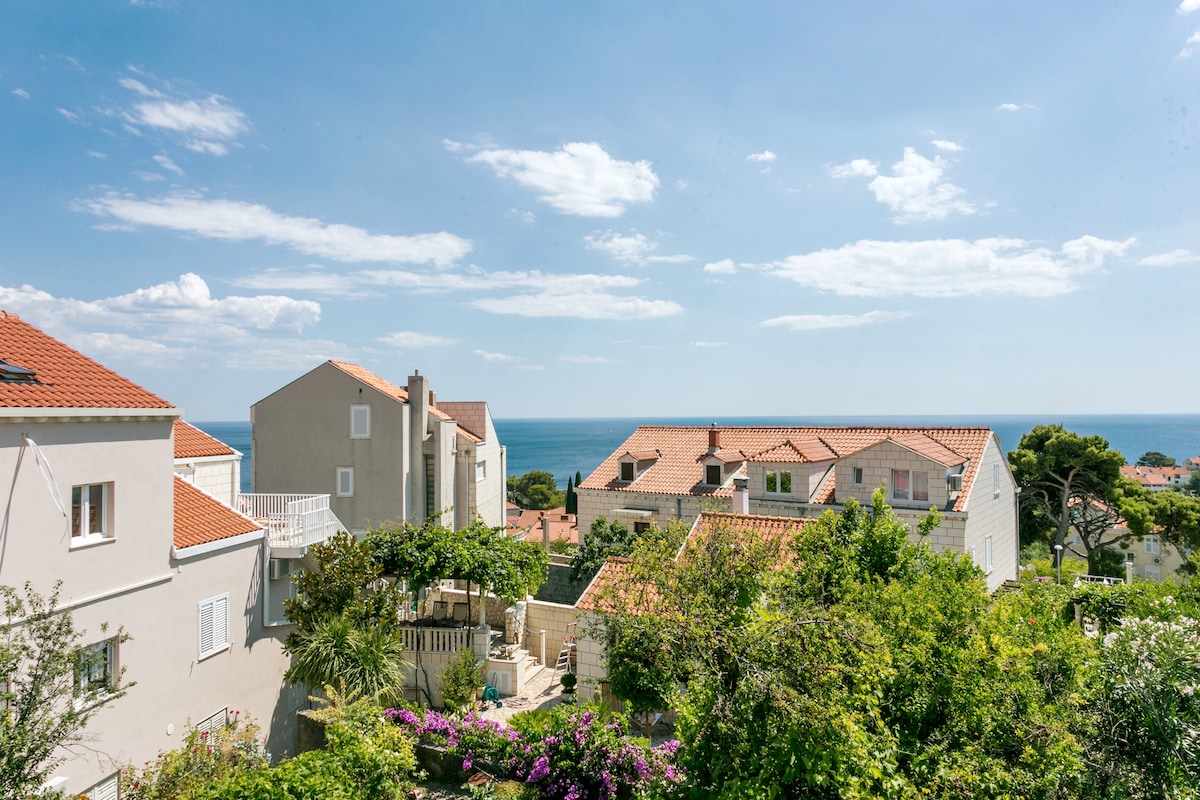 A scenic view encompasses homes with terracotta roofs and lush greenery, leading toward the sea. A clear blue sky stretches overhead, dotted with soft white clouds. The tranquil atmosphere highlights the natural beauty of the surrounding area.