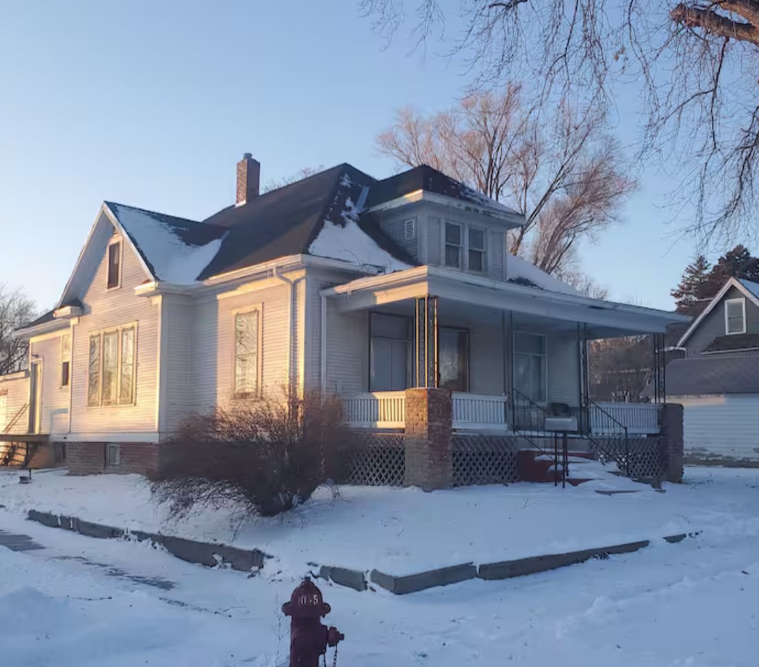 A classic two-story home is set against a snowy landscape, featuring a broad front porch with white railings. Sunlight highlights the architectural details of the house, including tall windows and a gabled roof. Sparse trees surround the property, enhancing the serene winter scene.