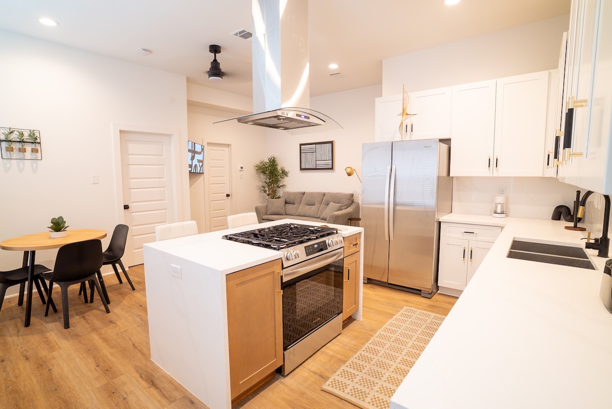 A modern kitchen is showcased, featuring a spacious island with a gas range and seating for four. Sleek stainless steel appliances and white cabinetry create an open-concept design. Natural light fills the area, highlighting the seamless flow into the adjacent living space.
