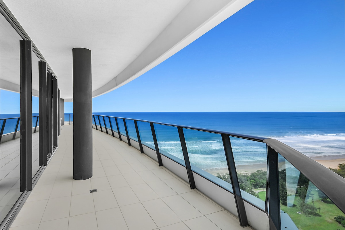 An expansive balcony offers panoramic oceanfront views, with clear blue skies above and sparkling waves below. The smooth tiled flooring contrasts with the sleek glass balcony railing, while columns support the curved overhang, creating a spacious and inviting outdoor area.