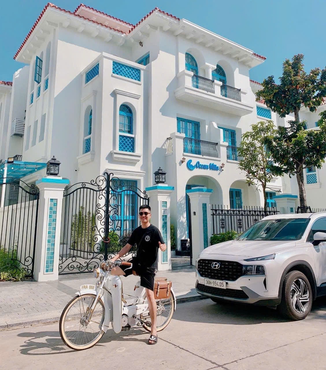 A modern villa features a bright white facade with ornate blue accents and large windows. A white vehicle is parked in front of the villa, while a bicycle in a light color leans against it. Lush greenery surrounds the property, providing a welcoming atmosphere.