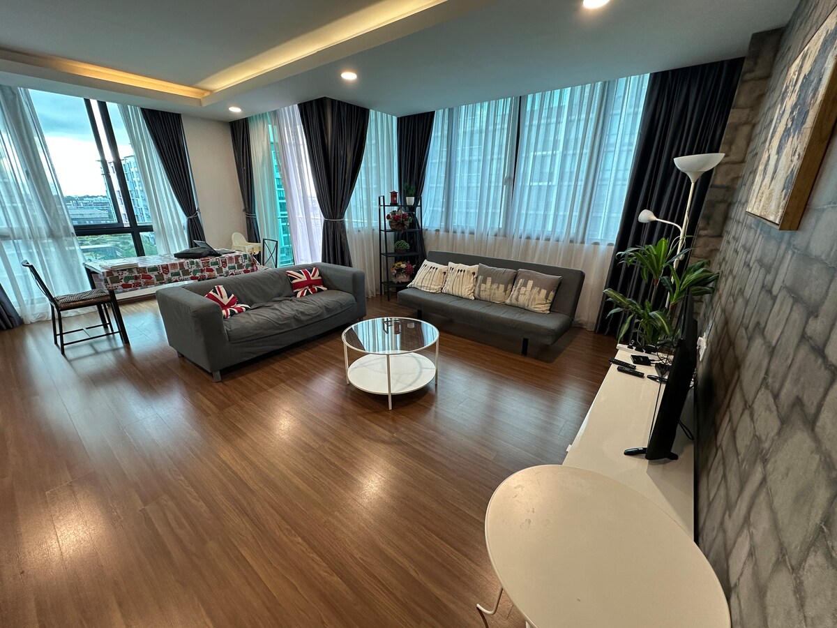 A spacious living area is filled with natural light from large windows dressed in sheer curtains. Two sofas with decorative cushions are positioned around a round coffee table, while potted plants and a neatly arranged shelf enhance the space. A flat-screen television is placed on an elegant console.
