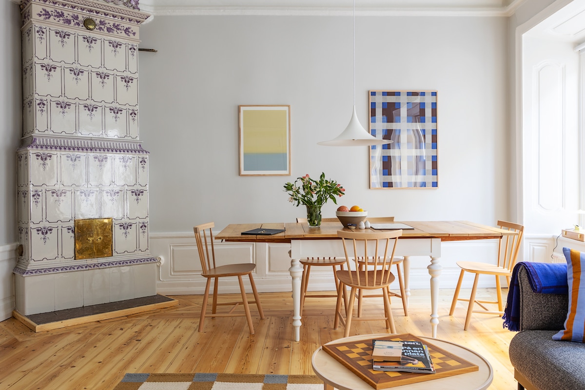 A stylish dining area features a wooden table surrounded by four light-colored chairs. A floral arrangement sits at the center of the table. A patterned rug lies beneath, and artwork adorns the walls, while a tiled fireplace adds a touch of charm to the space.