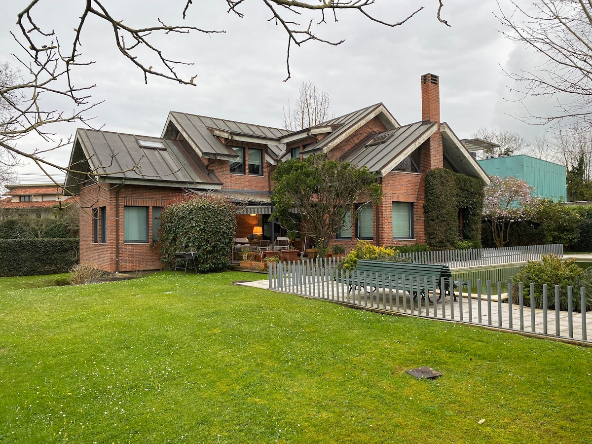 A spacious residential home is highlighted with a mix of brick and metal roofing. Surrounding greenery offers a manicured lawn with well-maintained bushes. The structure includes multiple roof angles, providing a unique architectural profile, while a patio area is visible in the front.