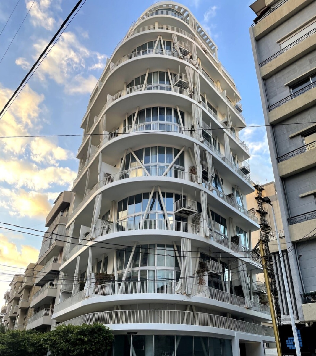 The image captures a modern, multi-story building featuring a unique architectural design with rounded edges and large glass windows. Balconies are visible along the exterior, adorned with plants. A clear sky with clouds adds depth to the scene.
