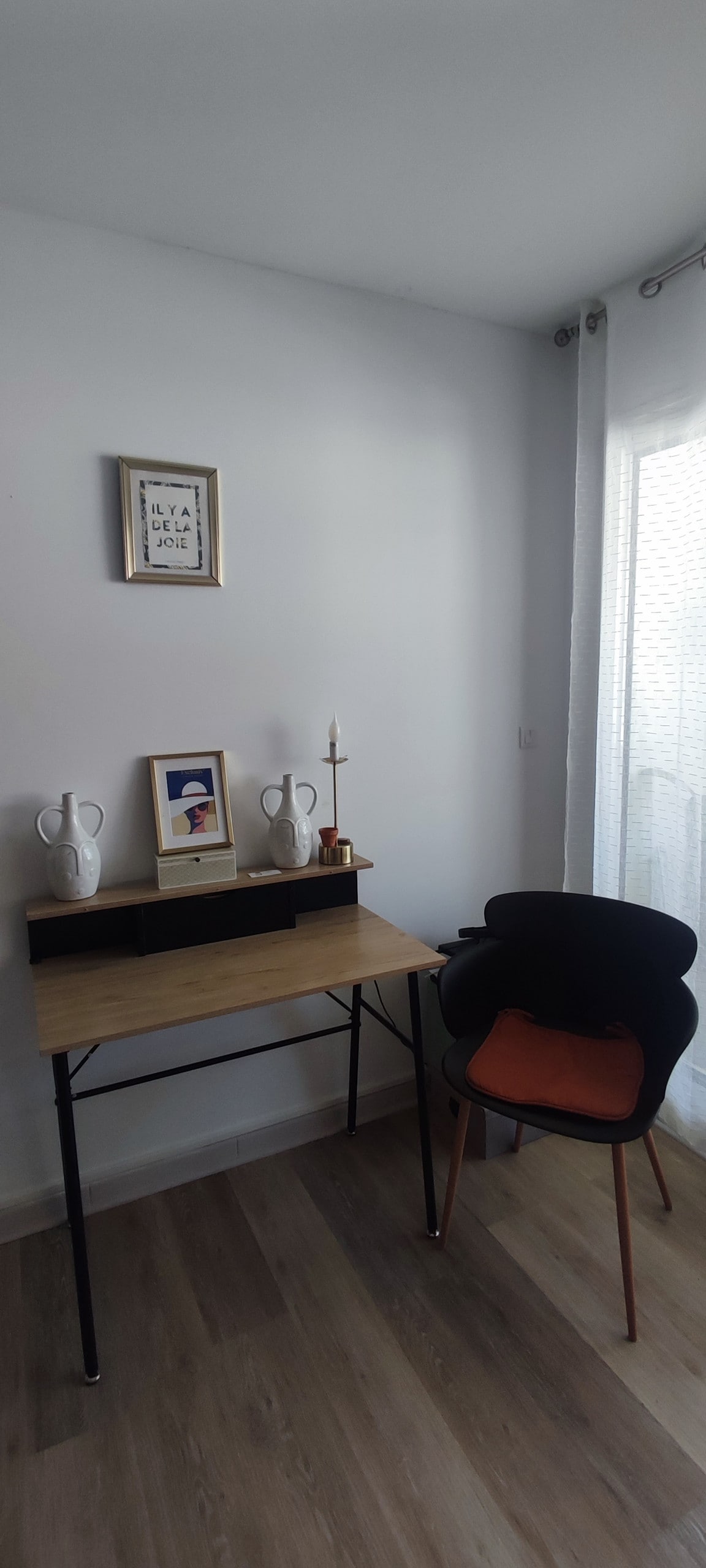 A functional workspace is presented, featuring a wooden desk with decorative items and a framed picture. A comfortable chair with an orange seat cushion is positioned next to the desk. Natural light enters through sheer curtains, illuminating the light-colored walls and flooring.