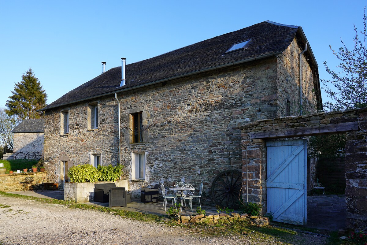 A restored stone barn is observed, featuring a rustic façade with a mix of narrow and wider windows. Outside, a patio area is visible, equipped with modern seating and a small dining set, surrounded by greenery, offering a serene outdoor space.