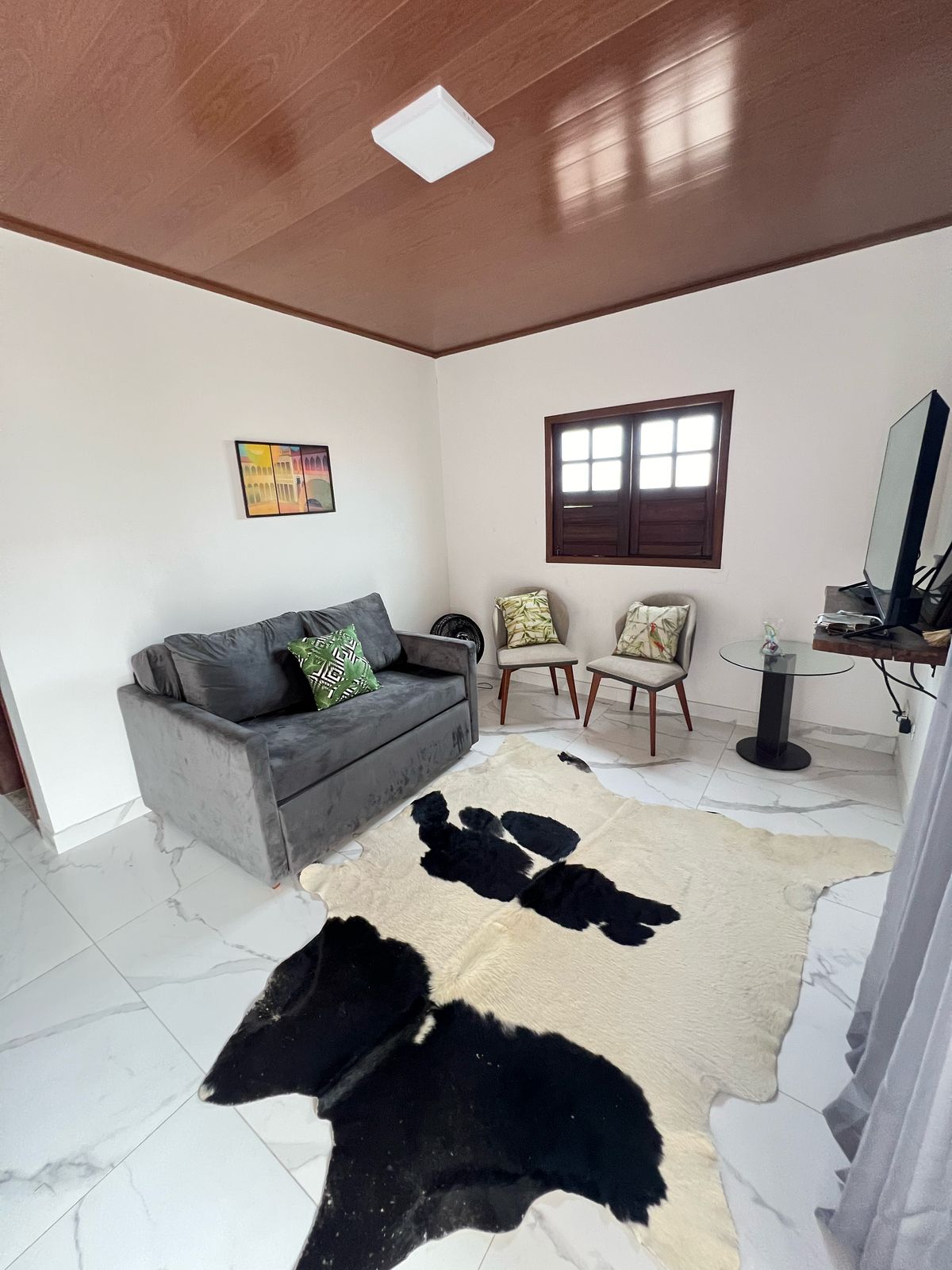 A cozy living area features a gray sofa, complemented by decorative pillows. Two wooden chairs are arranged beside a small glass table. A black and white rug lays on the marbled floor, while a window offers natural light, framed by brown wooden shutters.