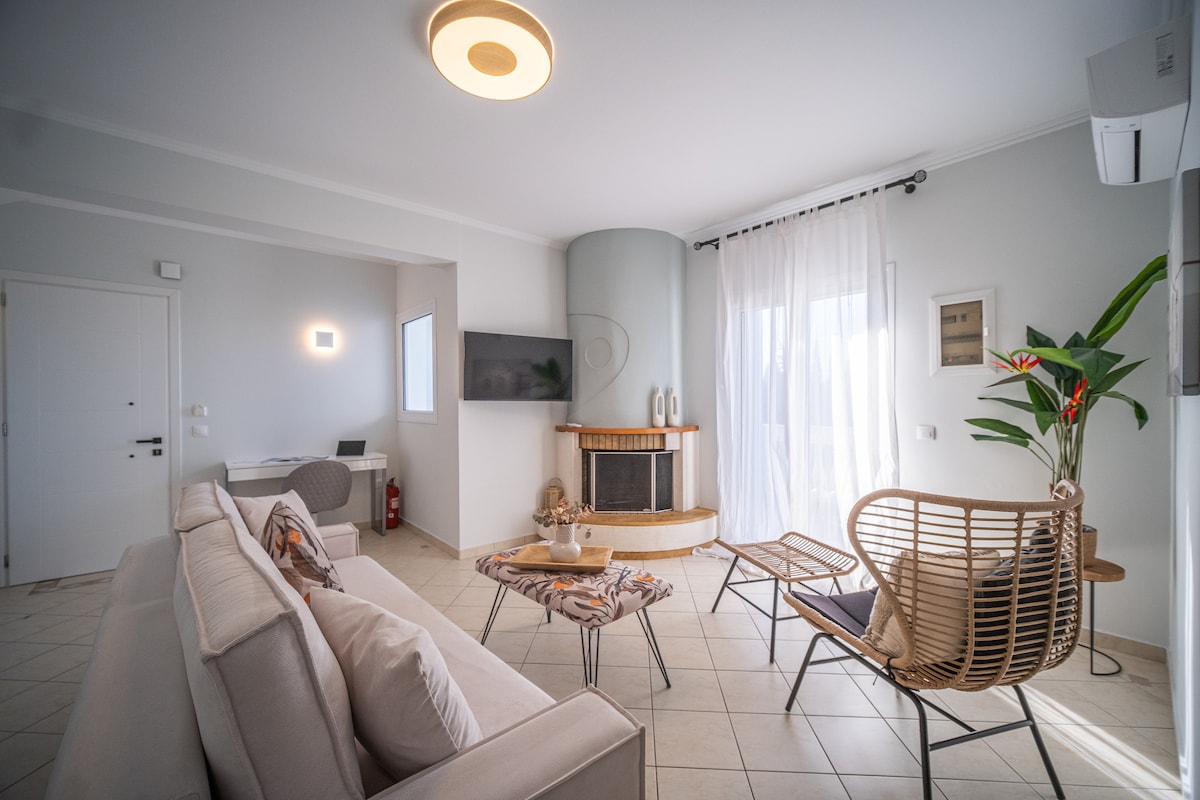 A spacious living area is furnished with a light-colored sofa and woven chairs. Natural light is allowed in through large windows, while a modern fireplace adds warmth. A flat-screen TV is mounted on the wall, and a potted plant enhances the tranquil setting.