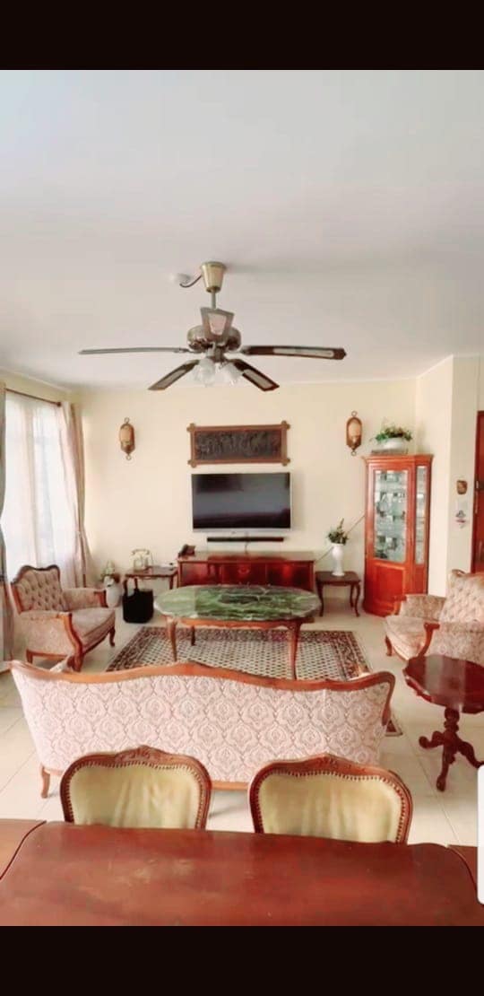 A welcoming living room is showcased, featuring comfortable seating arranged around a central area rug. A television is mounted on the wall, accompanied by decorative cabinets on either side. Natural light filters in through large windows with sheer curtains, enhancing the space's bright ambience.