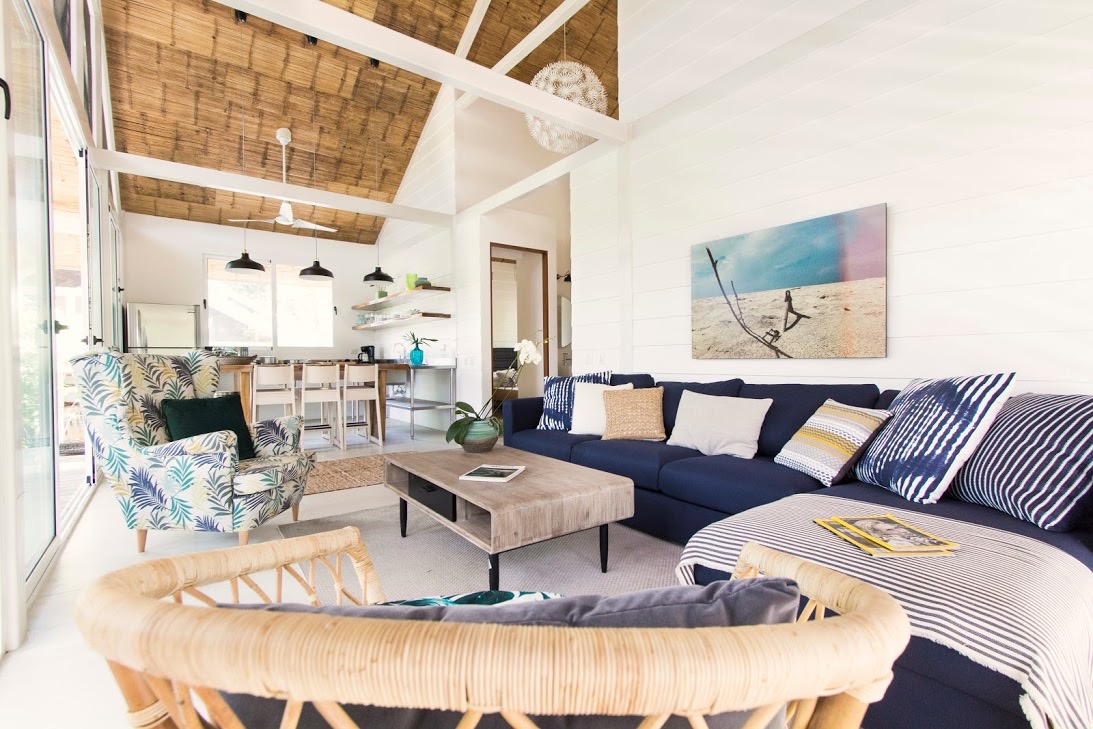 The bright living area showcases a spacious L-shaped couch adorned with various cushions, complemented by a light wooden coffee table. A vibrant armchair with floral patterns is nearby. Large sliding glass doors allow natural light to fill the space, revealing views of the kitchen and dining area in the background.