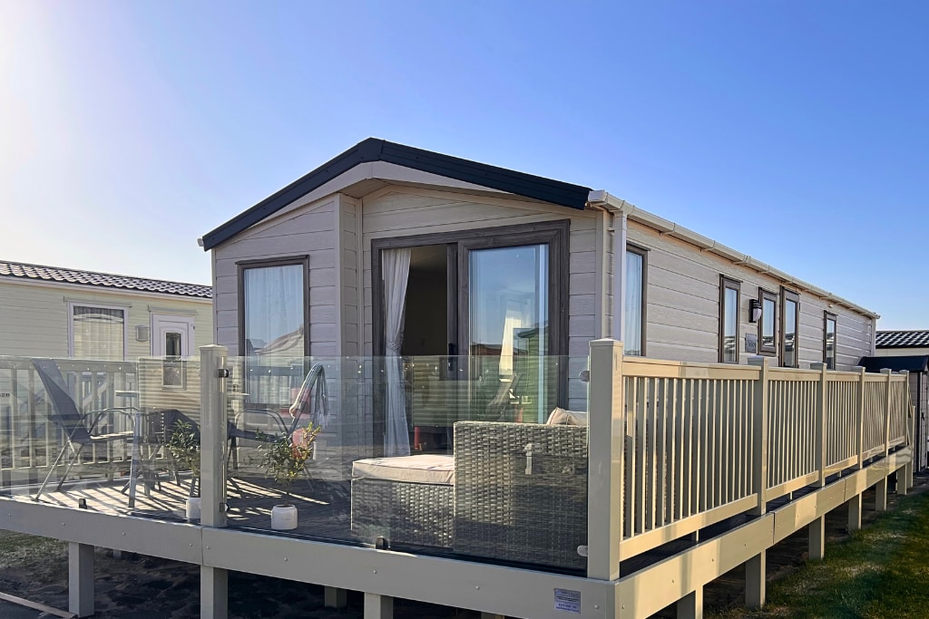 A charming caravan is displayed, featuring wrap-around decking with glass panels. Outdoor seating arrangements, including a sofa and lounge chairs, can be seen. Large windows reflect natural light, enhancing the spacious interior view. The structure is set against a clear blue sky.