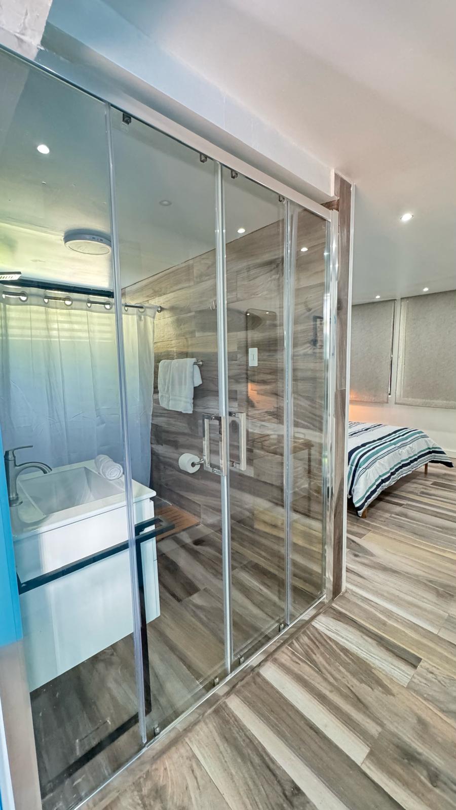 A modern bathroom features a sleek glass shower enclosure alongside a stylish freestanding tub. Contemporary fixtures are present, complemented by wooden accents on the walls and floor. In the background, the inviting sleeping area is visible, illuminated by soft lighting.
