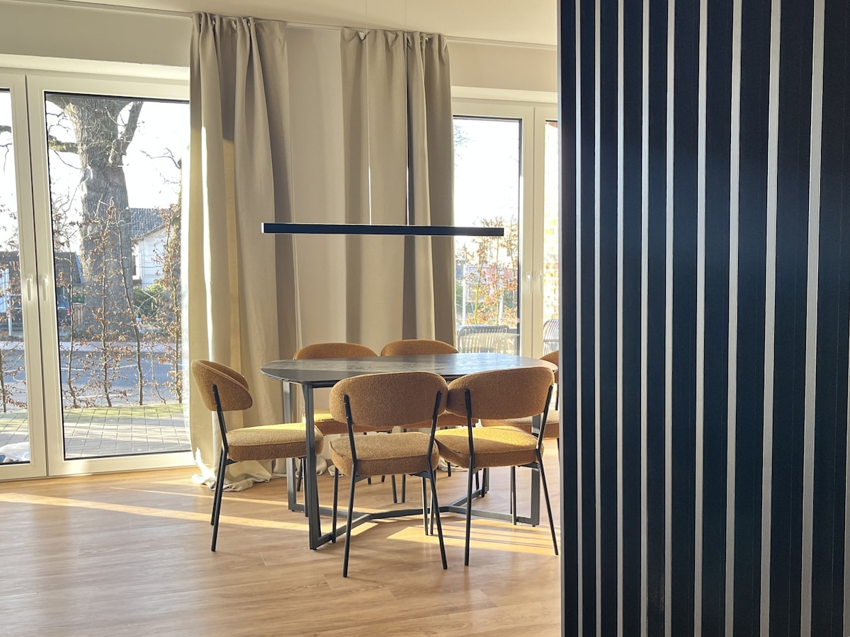A dining area featuring a round table surrounded by six chairs is illuminated by natural light from large floor-to-ceiling windows. Elegant curtains frame the windows, and modern lighting hangs above the table, creating a conducive environment for meals and gatherings.