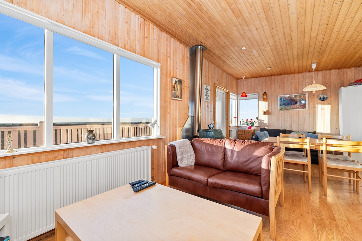 The living area features warm wooden walls and a light wooden ceiling, creating a cozy atmosphere. A comfortable brown sofa faces large windows, providing expansive views. A coffee table sits in front of the sofa, with a dining area visible in the background, complemented by a decorative pendant light.
