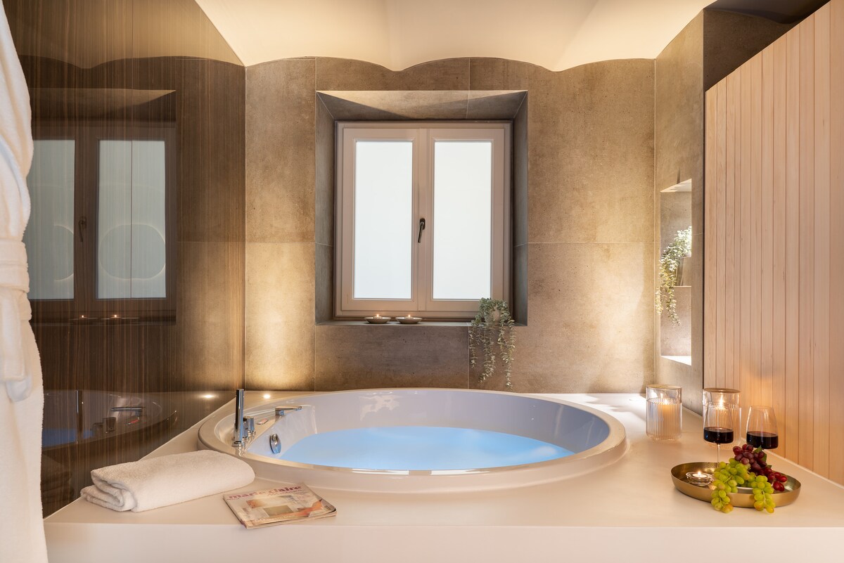 A spacious bathroom features a circular soaking tub surrounded by soft lighting. A window provides natural light, while a plush bathrobe is hung nearby. Fresh fruit and candles on the counter add a touch of relaxation, creating an inviting space for unwinding.