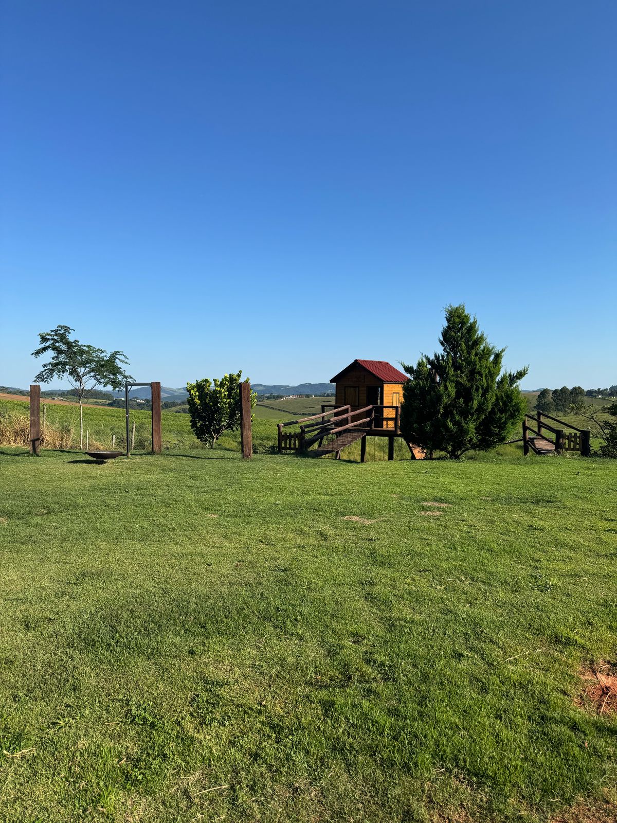 A spacious grassy area is complemented by a wooden playhouse with a red roof and stairs. Surrounding greenery includes trees and shrubs, while a wooden fence lines the perimeter. A clear blue sky provides a bright backdrop, enhancing the natural setting.