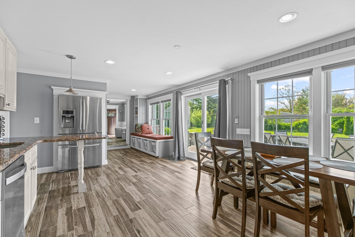 A bright, open kitchen and dining area features light-colored cabinetry and a spacious island. Large windows provide ample natural light, showcasing a view of the patio and greenery beyond. A comfortable dining table is positioned beside rustic wooden chairs, enhancing the inviting atmosphere.
