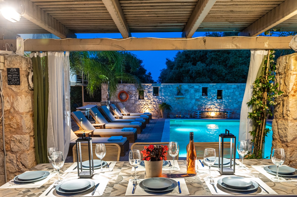 An outdoor dining area is presented with a stone table set for several guests. The table is adorned with tableware, glasses, and a small potted plant. In the background, a private pool is surrounded by sun loungers under soft evening lighting, framed by lush greenery.