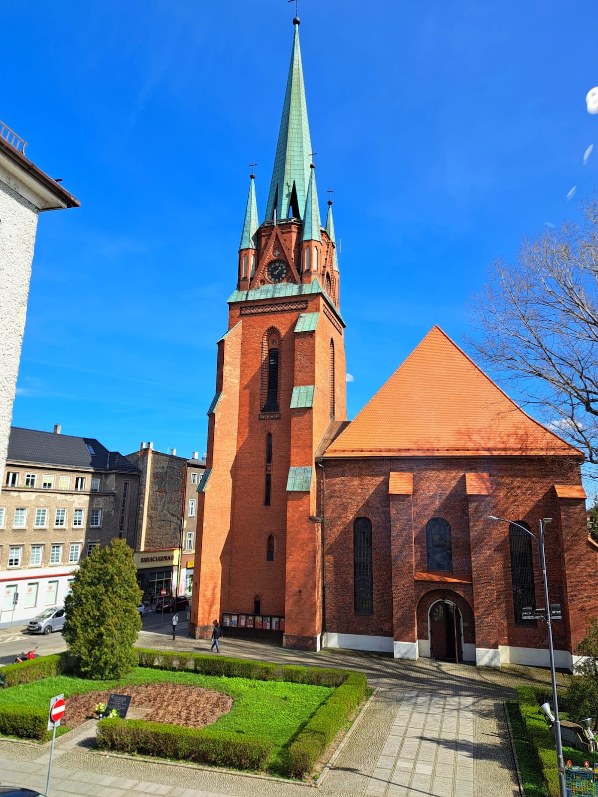 A tall, pointed tower with a green spire rises above a red brick church. The structure features distinct Gothic architectural elements and is surrounded by well-maintained greenery, including a small garden area. Clear blue skies provide a bright backdrop.
