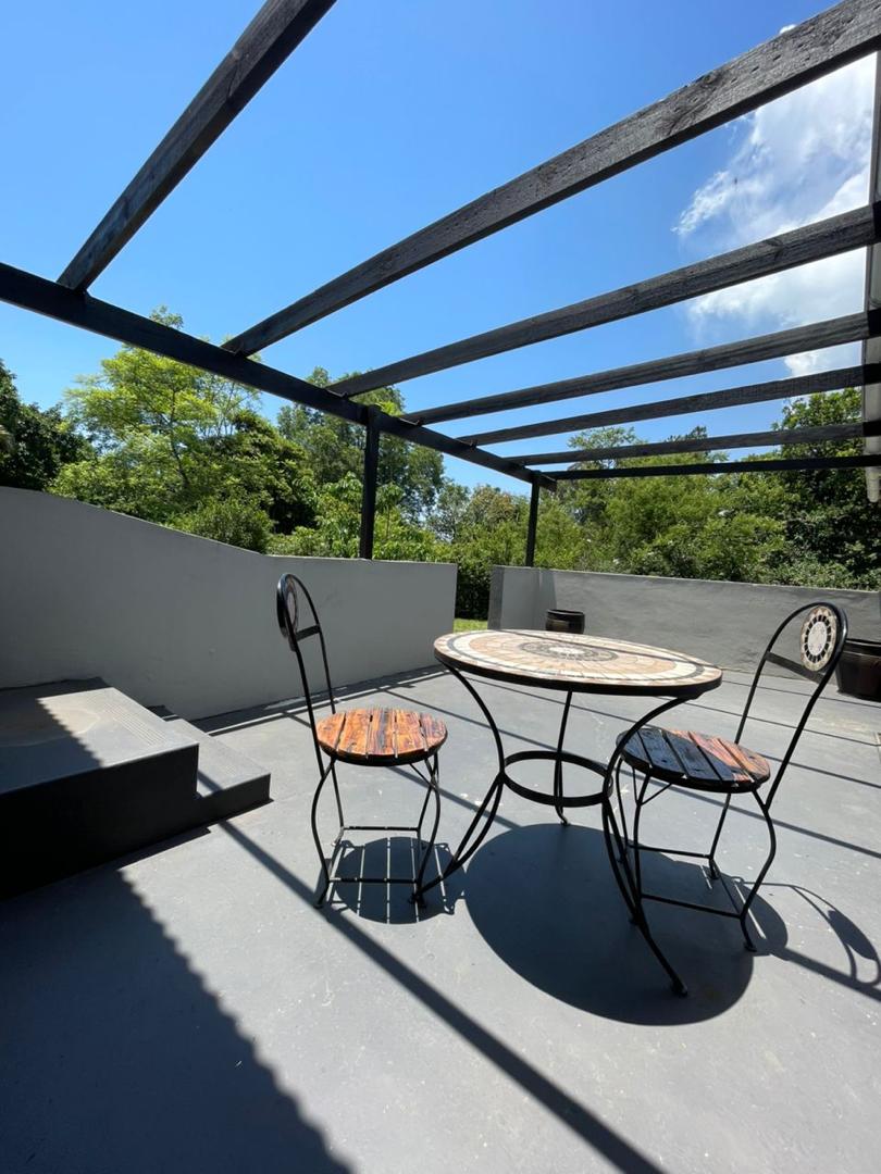 An outdoor seating area features a round table accompanied by two black metal chairs. A partially covered patio allows for natural light to stream in, while lush greenery is visible in the background. The structure's beams create an open, airy atmosphere.