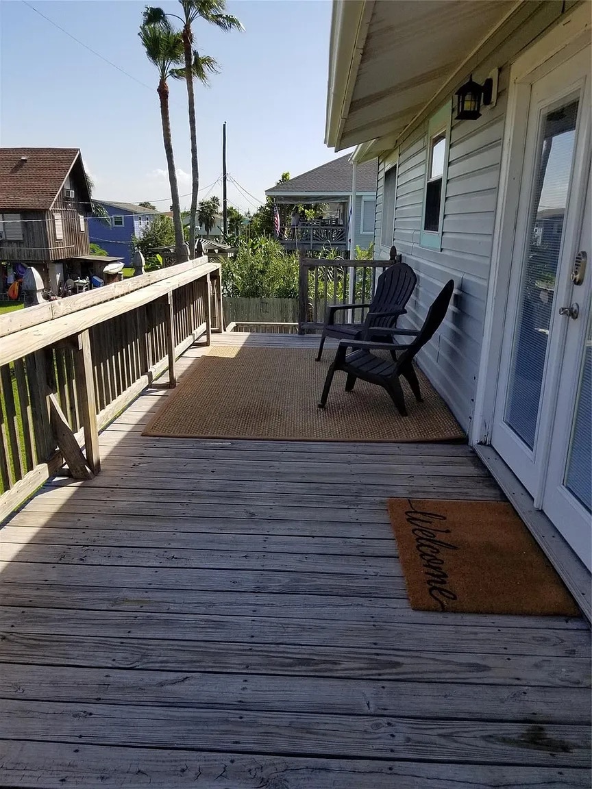 A wooden deck is presented, featuring two black chairs positioned on a beige mat. A welcome mat greets visitors at the entrance. Lush greenery and palm trees are visible in the background, and a clear sky creates a bright atmosphere.
