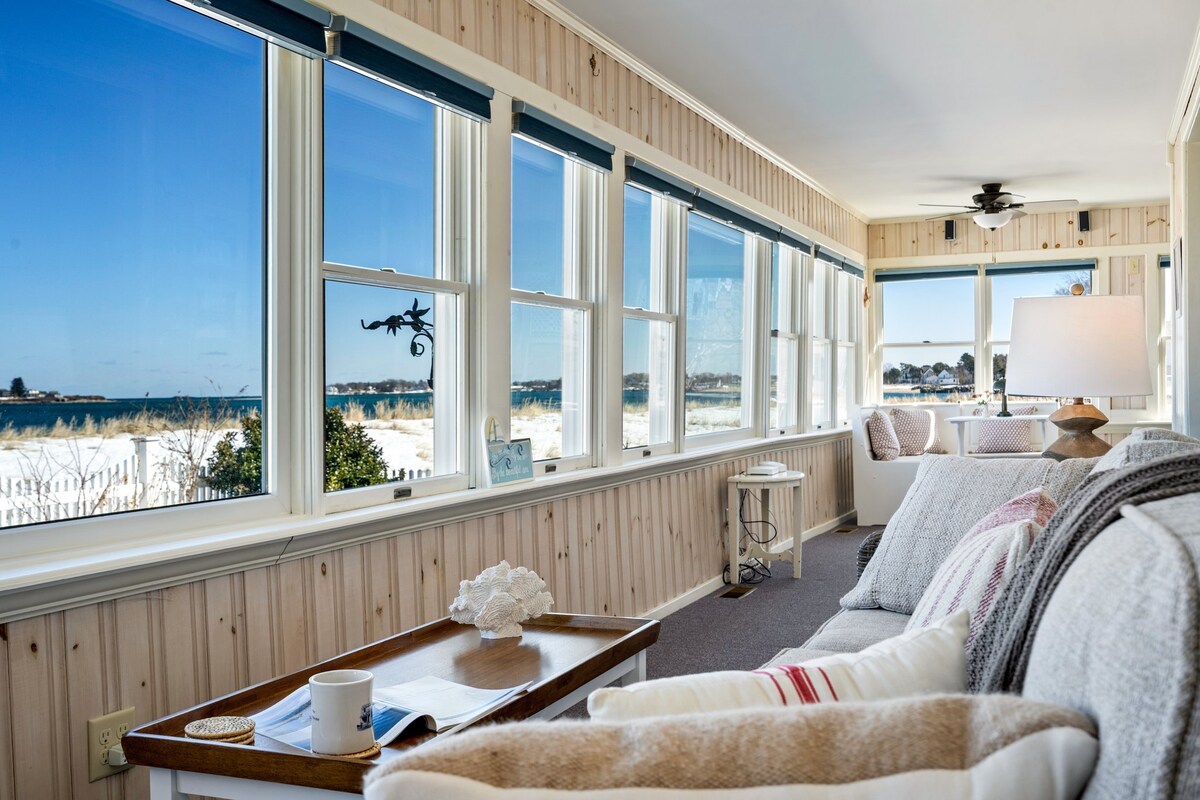 A sunlit enclosed porch features expansive windows that provide views of the beach and dunes. A light-colored sofa with plush cushions is positioned near a small table, enhancing the relaxed coastal feel of the space. Soft decor elements contribute to a serene environment.