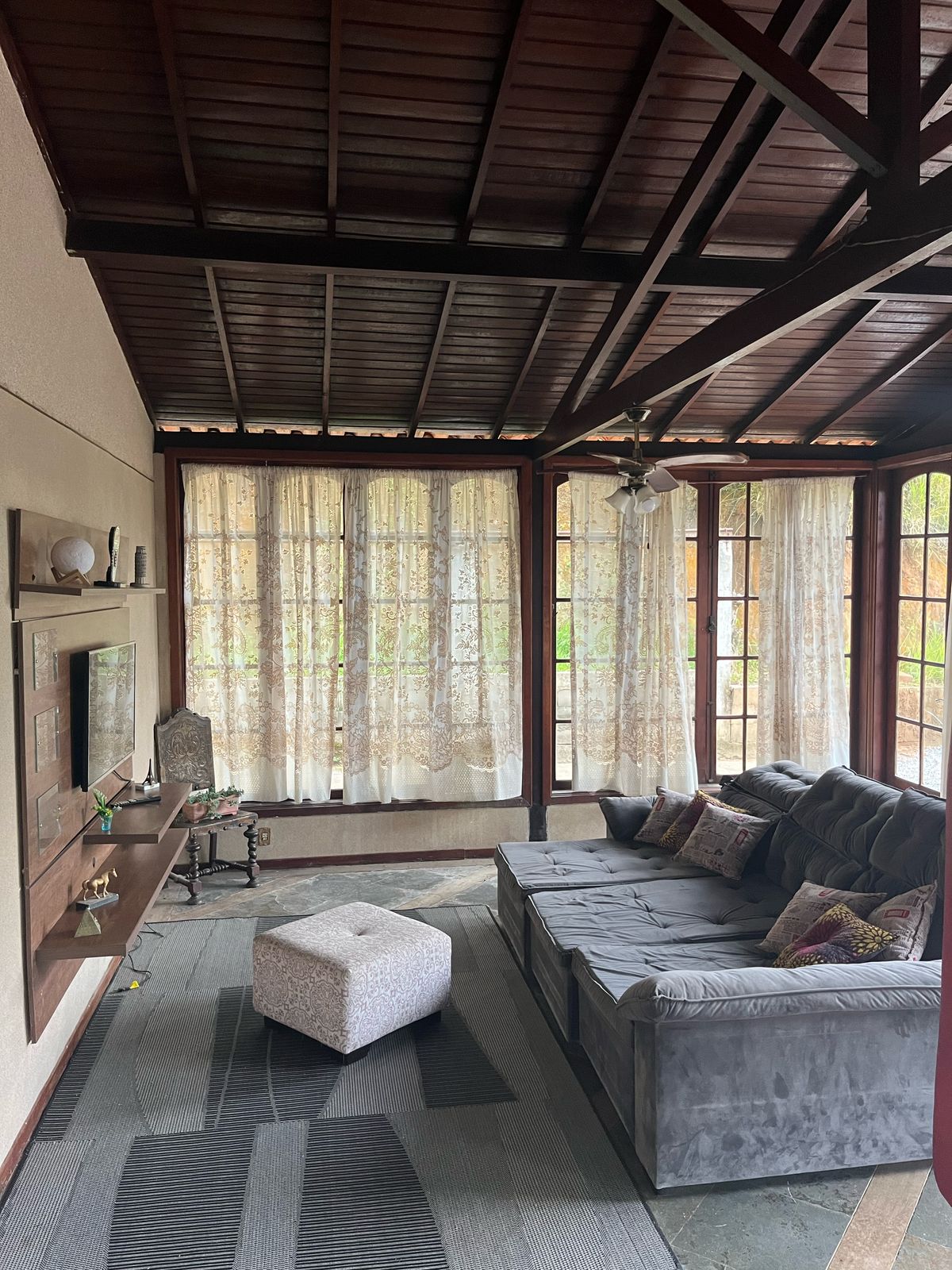A spacious living area features large windows draped with light, airy curtains, allowing natural light to fill the room. A comfortable sectional sofa is positioned around a plush ottoman, with decorative pillows adorning the seating. A wooden shelf displays decorative items and a television is mounted on the wall.