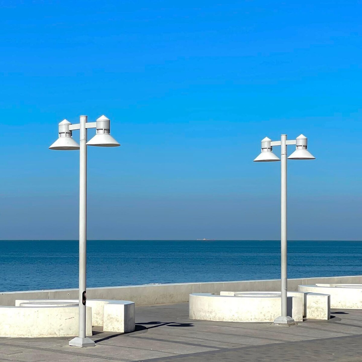 Two modern light poles stand along a beachfront walkway, casting soft shadows on the tiled surface. The ocean stretches out in the background, meeting a clear blue sky. The horizon is visible, creating a serene coastal atmosphere.