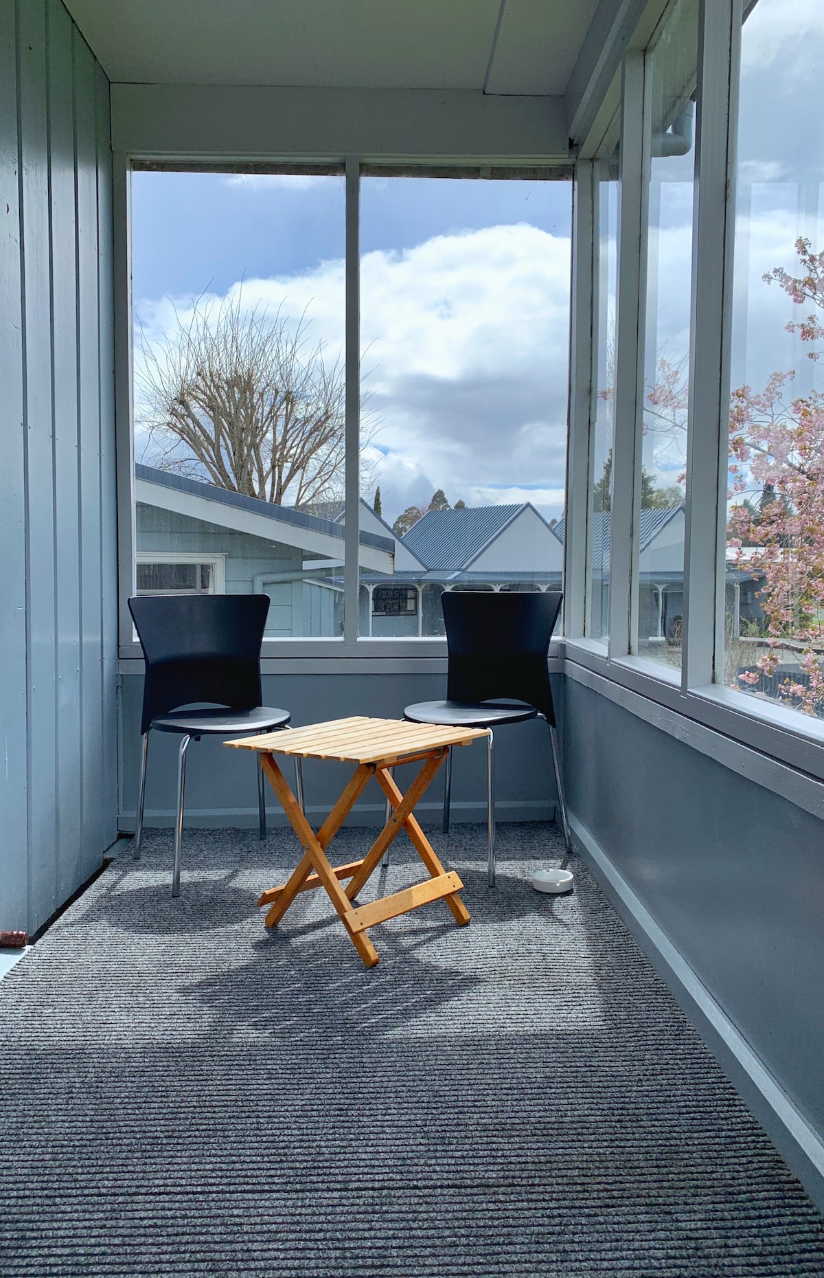 A covered outdoor area features two chairs positioned around a small, foldable table. Large windows allow natural light to fill the space, while a gray carpet adds warmth. The view includes neighboring buildings and trees, creating a serene environment.