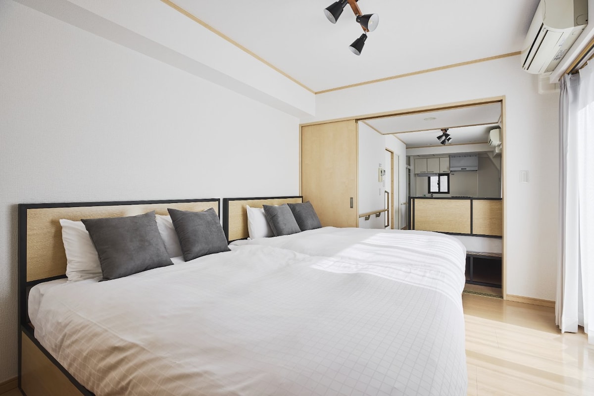 A well-lit bedroom is furnished with two spacious beds, each adorned with neutral-colored linens and pillows. Natural light filters in through a sheer curtain, while a wooden partition leads to a cozy living area visible beyond.