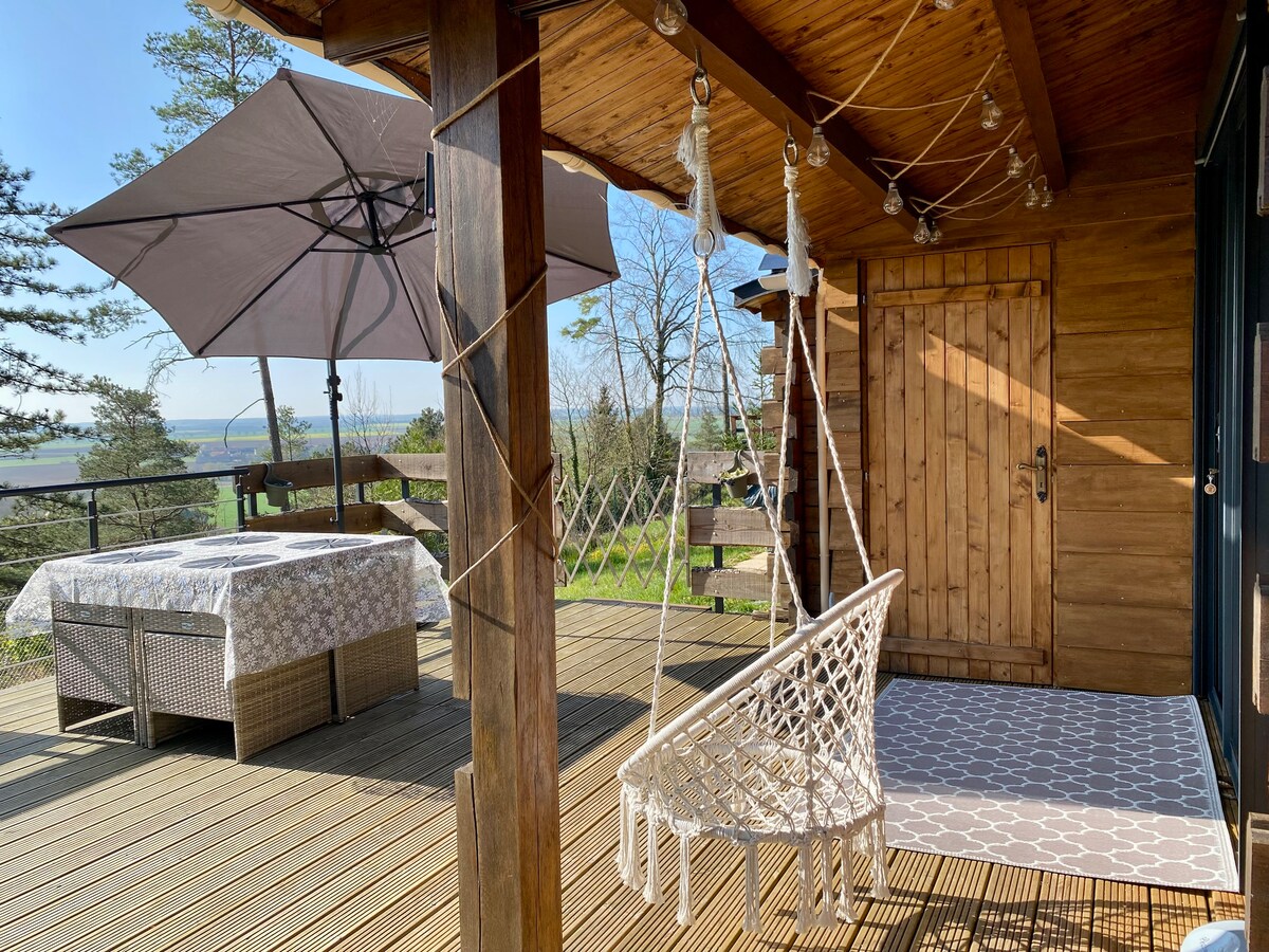 A wooden terrace is featured, showcasing a comfortable seating area covered by a large umbrella. A cozy swing chair is suspended from the porch roof, with a decorative rug placed on the wooden flooring. Natural light highlights the surrounding trees and landscape.