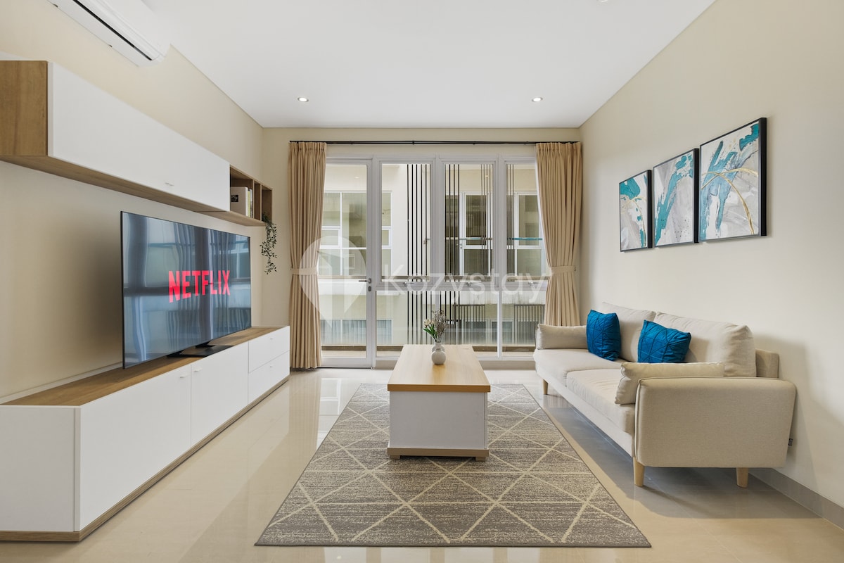 A spacious living area features a large TV displaying Netflix. A light-colored sofa, accompanied by decorative blue cushions, is positioned near a natural light-filled window. A simple coffee table rests on a geometric-patterned rug, enhancing the modern design of the space.