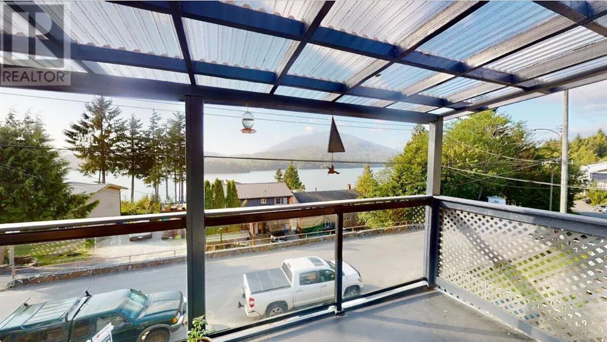 A covered balcony offers expansive views of the surrounding landscape, including a body of water and distant mountains. A sleek railing surrounds the area, with a few decorative items visible. The clear sky above suggests a bright day, enhancing the serene setting.