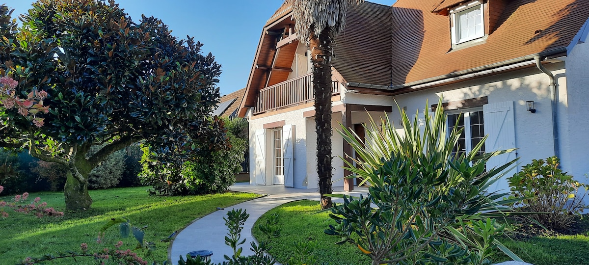 The exterior of the villa features a well-manicured garden with various greenery, including palm and flowering plants. A curved walkway leads to the welcoming entrance, where large glass doors provide a view into the interior. The house is complemented by a sloped roof and a balcony.