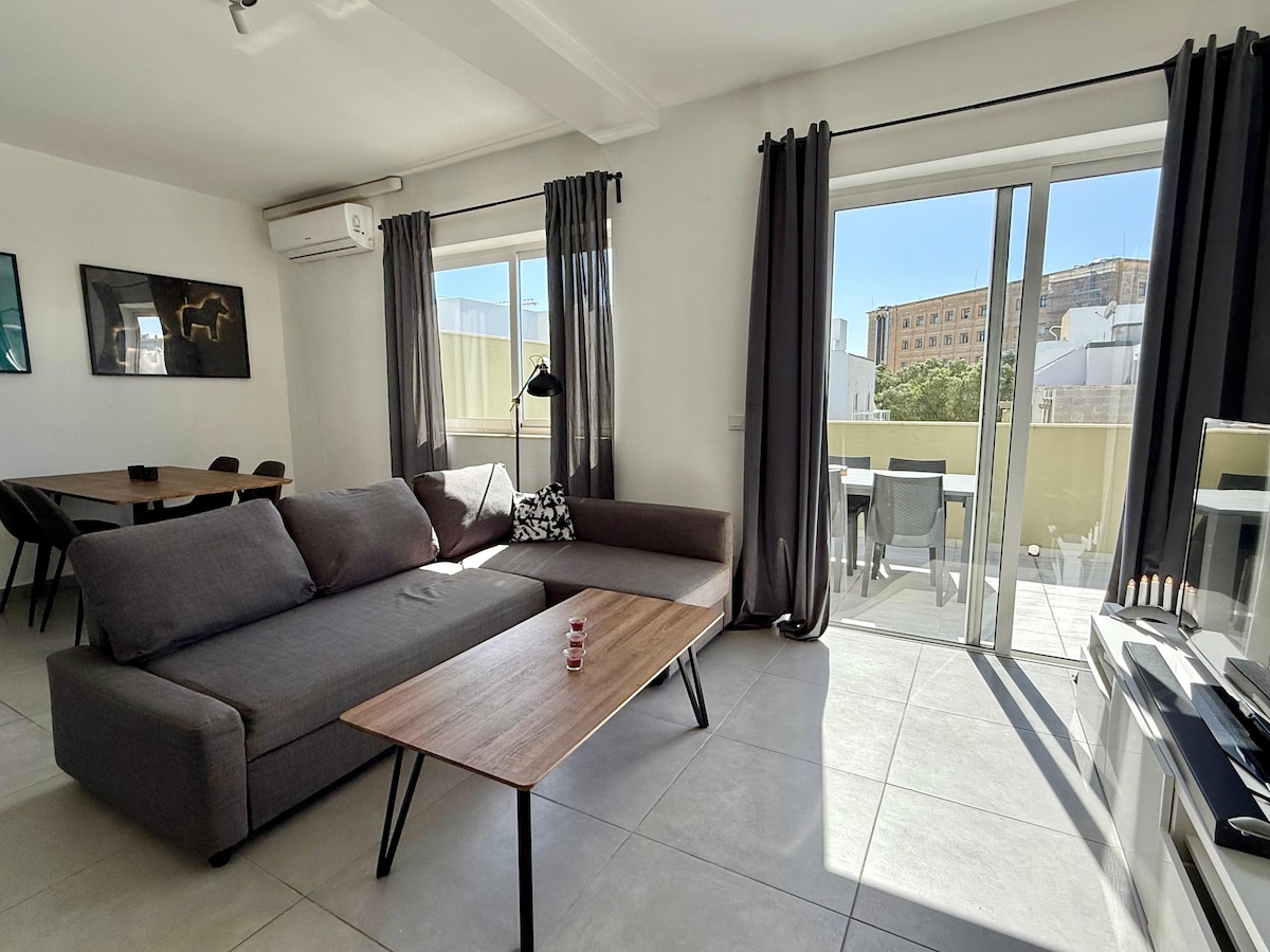 A spacious open-plan living area features a gray sectional sofa and a wooden coffee table. Large glass doors provide access to a terrace, allowing natural light to fill the room. A dining table is visible in the background, complemented by stylish wall art.
