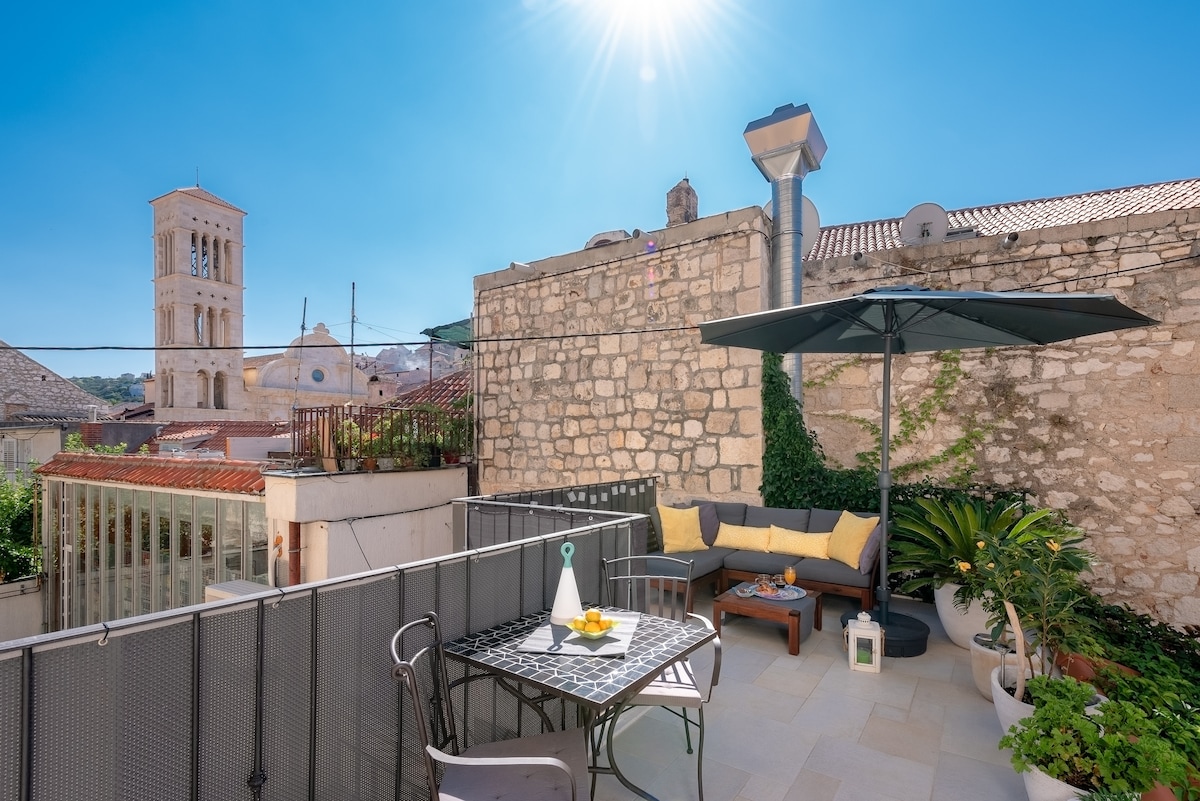 A terrace is displayed featuring a cozy seating area with cushions and a table set for two. An umbrella provides shade, and flowering plants add a touch of greenery. In the background, a historic tower stands against a clear blue sky.