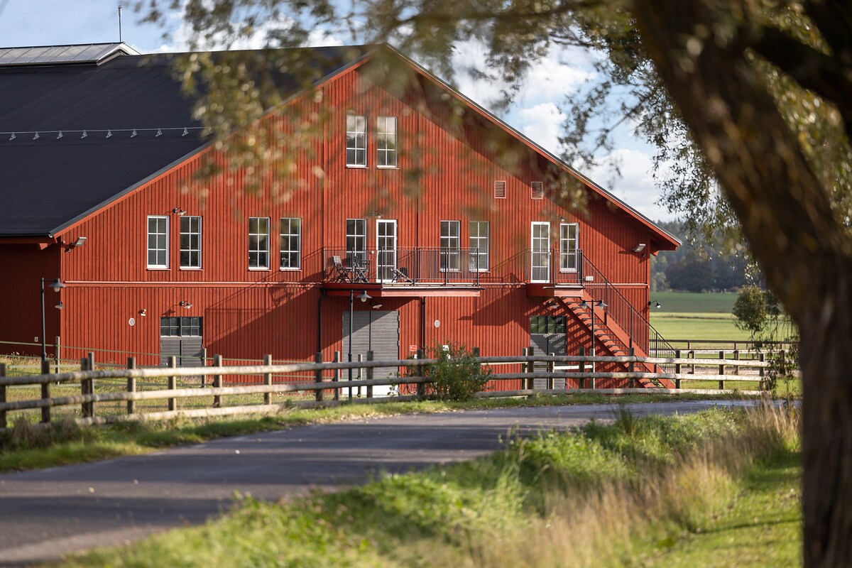 A large red barn-style building is set against a backdrop of open fields. Multiple windows welcome natural light, and a staircase leads to an upper entrance. A wooden fence borders a pathway, providing a sense of serenity in the surrounding landscape.