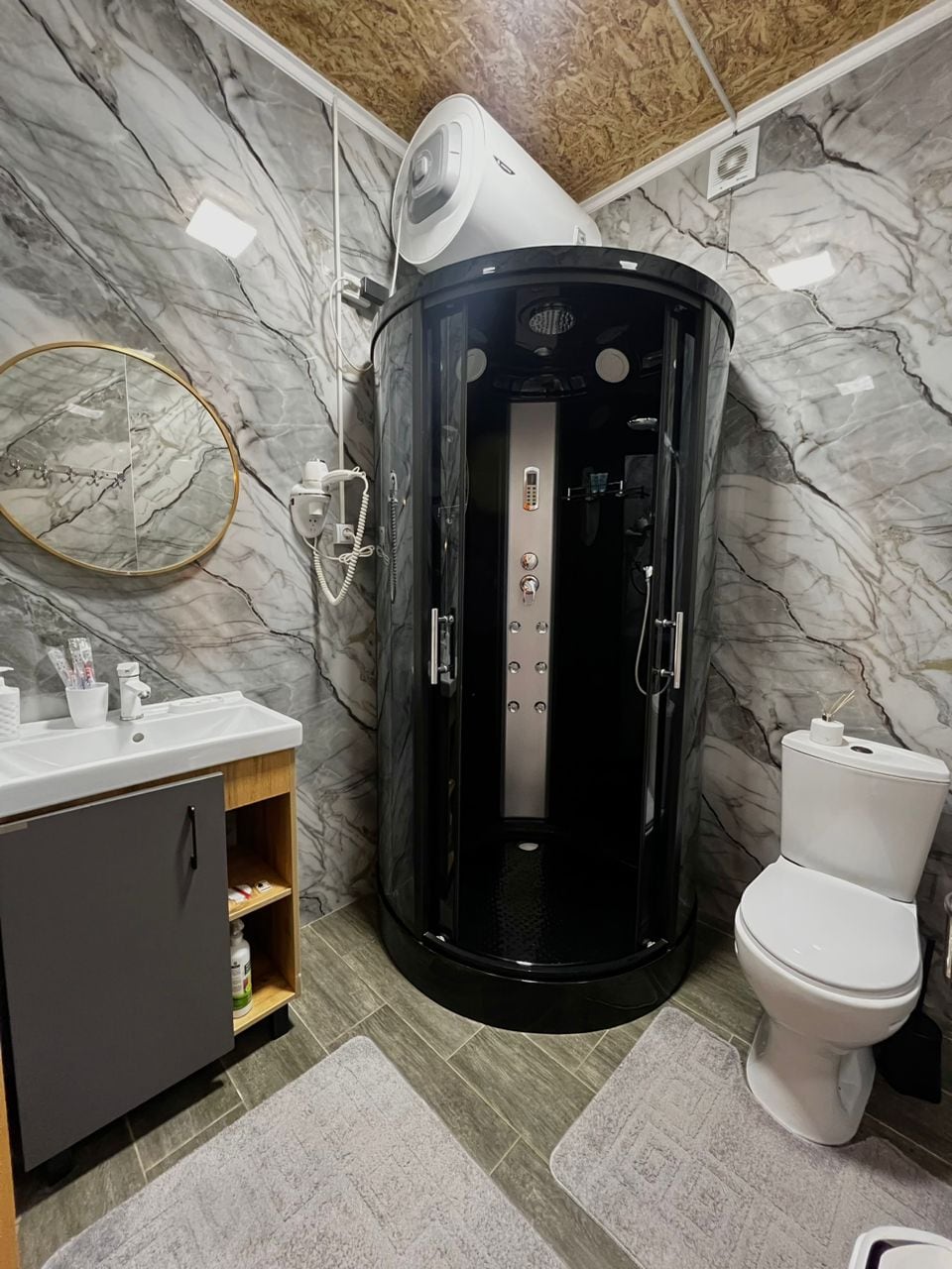 A modern bathroom features a sleek black shower unit, equipped with multiple settings. Natural stone walls add texture, while a round mirror complements the space. A countertop with a sink is situated next to a stylish cabinet, and a toilet is positioned against the wall.