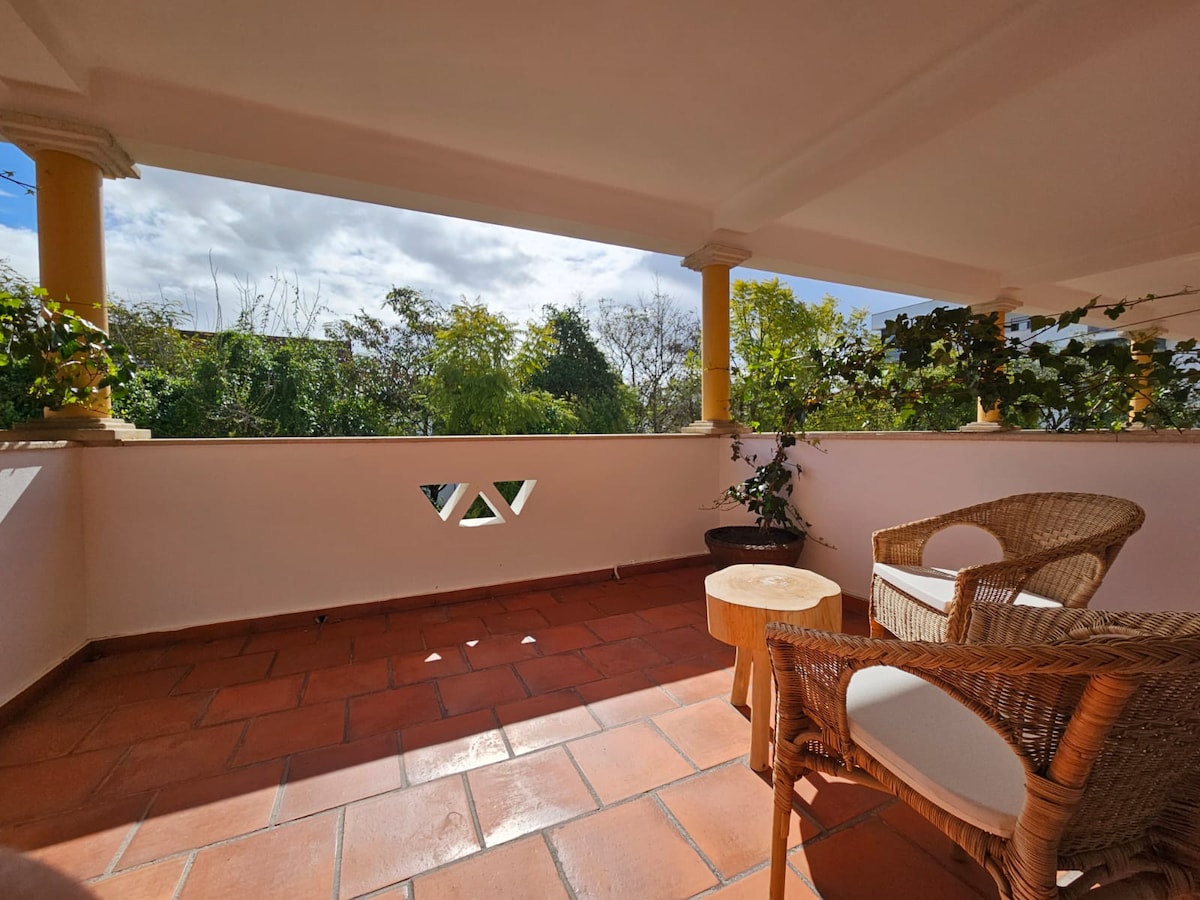 A terrace area is featured, showcasing terracotta tiles and two woven chairs positioned around a small wooden table. Lush greenery is visible beyond the railing, with patches of blue sky and clouds creating a serene outdoor atmosphere.