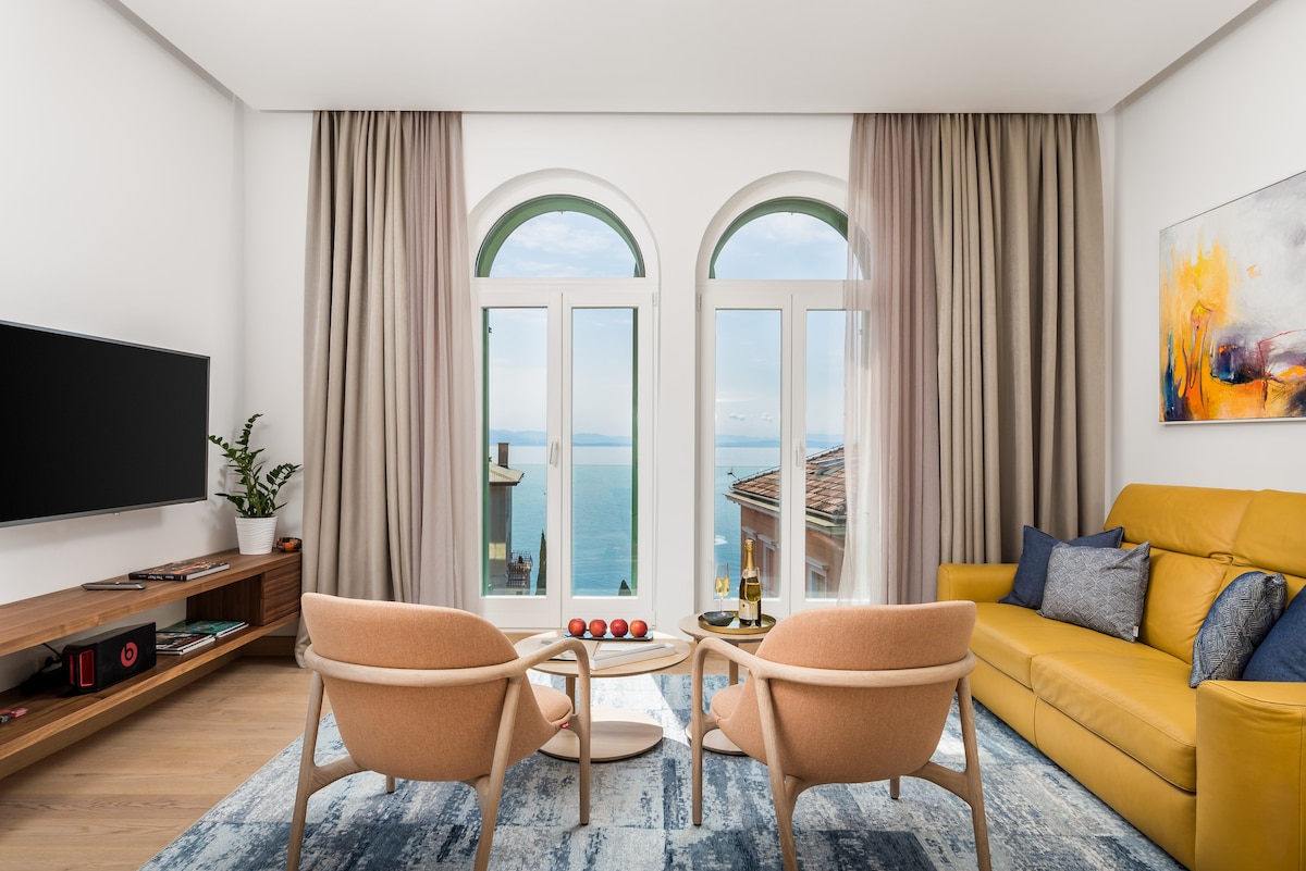 A bright living area features large arched windows that provide a view of the sea. Two beige armchairs are positioned around a small coffee table, while a yellow sofa adds a pop of color. A flat-screen TV is mounted on a wooden console, complemented by elegant drapes.