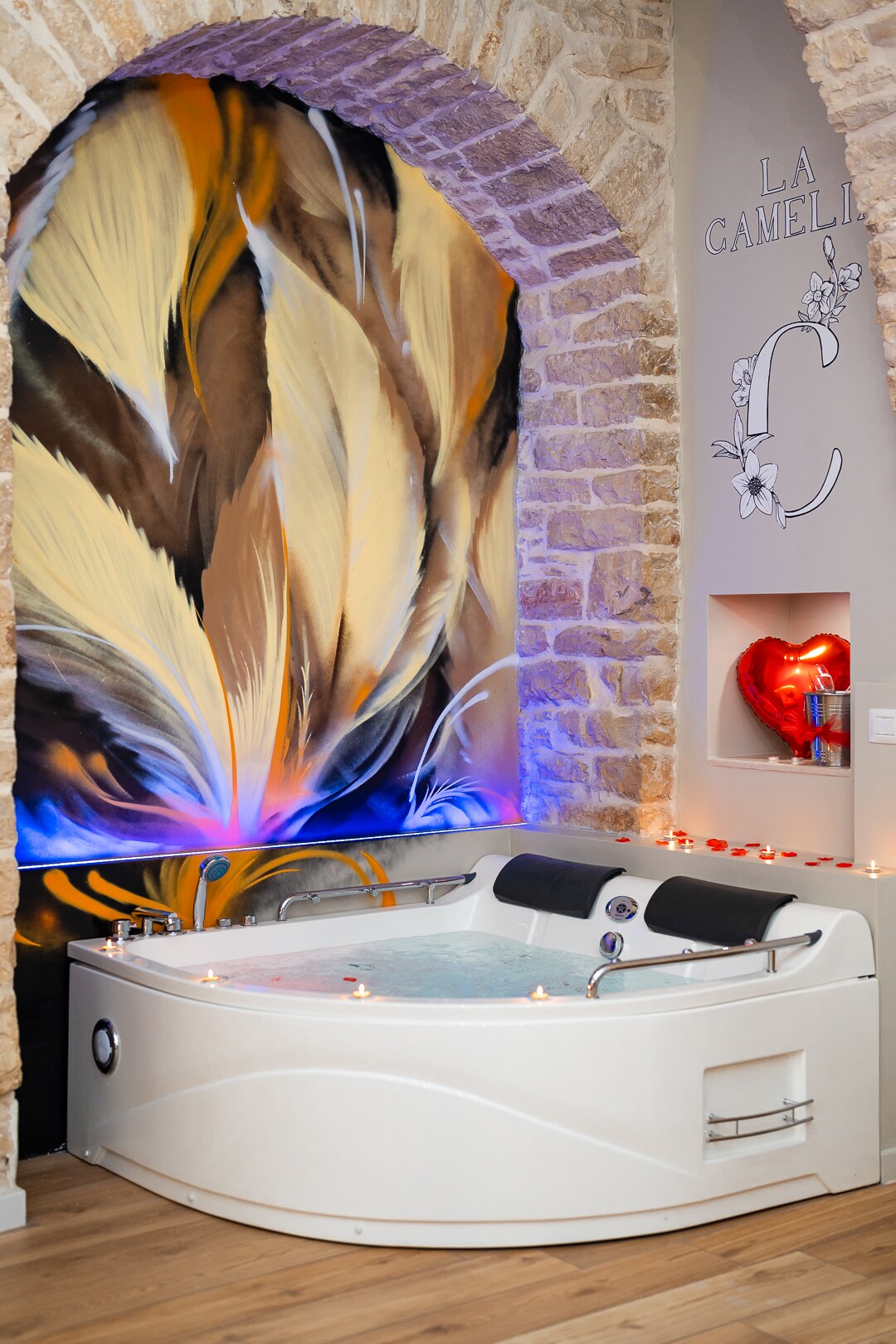 A spacious hot tub is positioned against a stone wall, accented by an artistic mural featuring soft, flowing colors. Soft lighting creates a serene ambiance, while decorative candles are arranged around the tub, inviting relaxation. A modern design is complemented by comfortable seating along the edge.