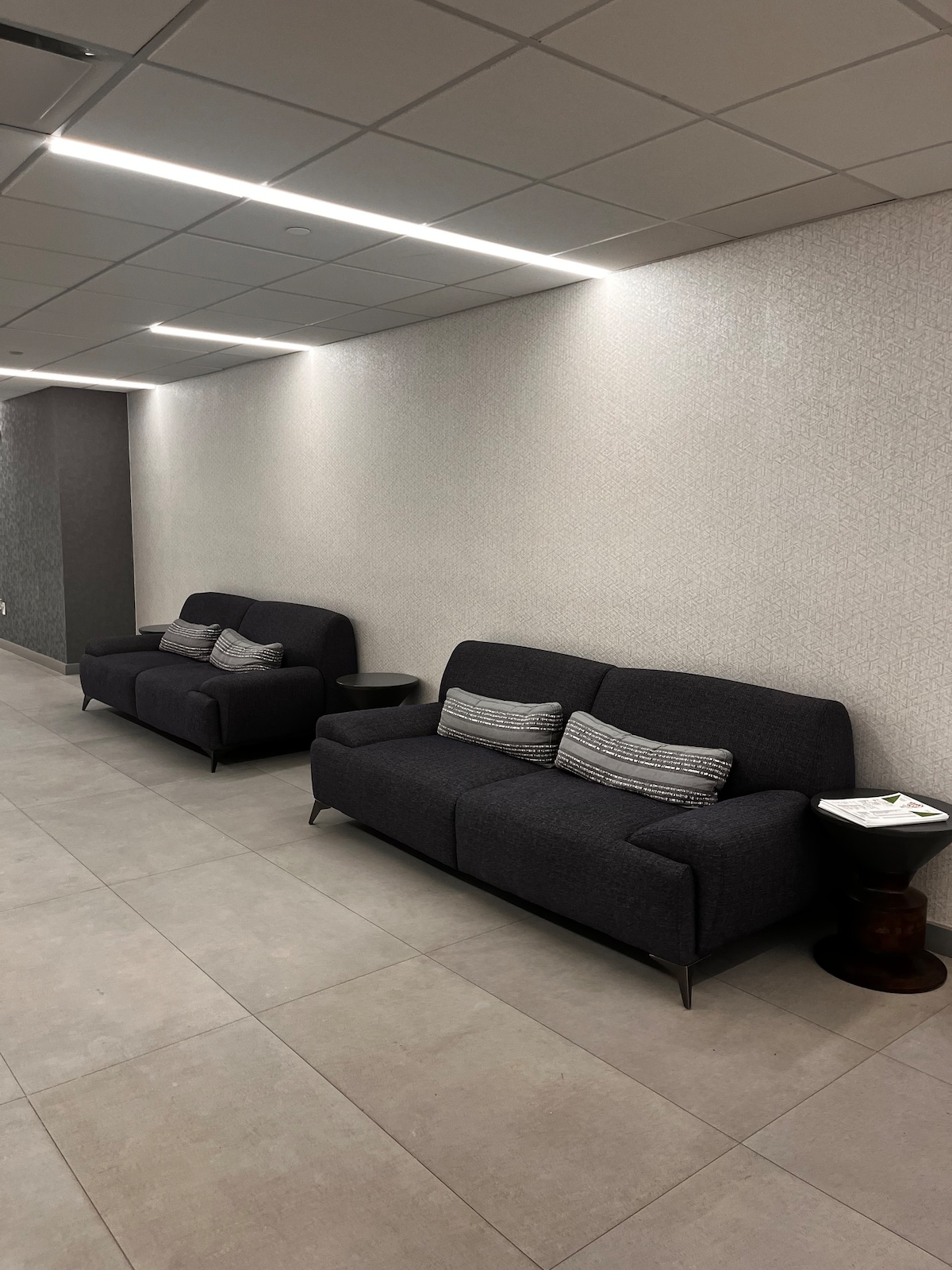 Two modern sofas in a neutral-toned lounge area are paired with small side tables. Soft lighting highlights the textured wall, contributing to a comfortable atmosphere for relaxation or casual gatherings.