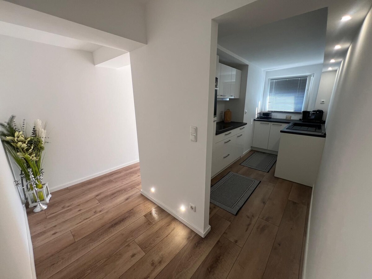 The modern kitchen is framed by light-colored cabinetry and features a countertop with kitchen essentials. Soft lighting illuminates the space, highlighting the clean lines and minimalistic decor. A hallway leading to the kitchen is visible, showcasing wood flooring and a decorative plant arrangement.